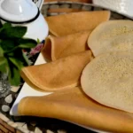 A serving plate with several round Moroccan pancakes, accompanied by a teapot and fresh mint leaves on a decorative tray