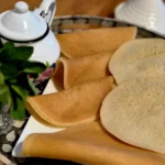 A serving plate with several round Moroccan pancakes, accompanied by a teapot and fresh mint leaves on a decorative tray