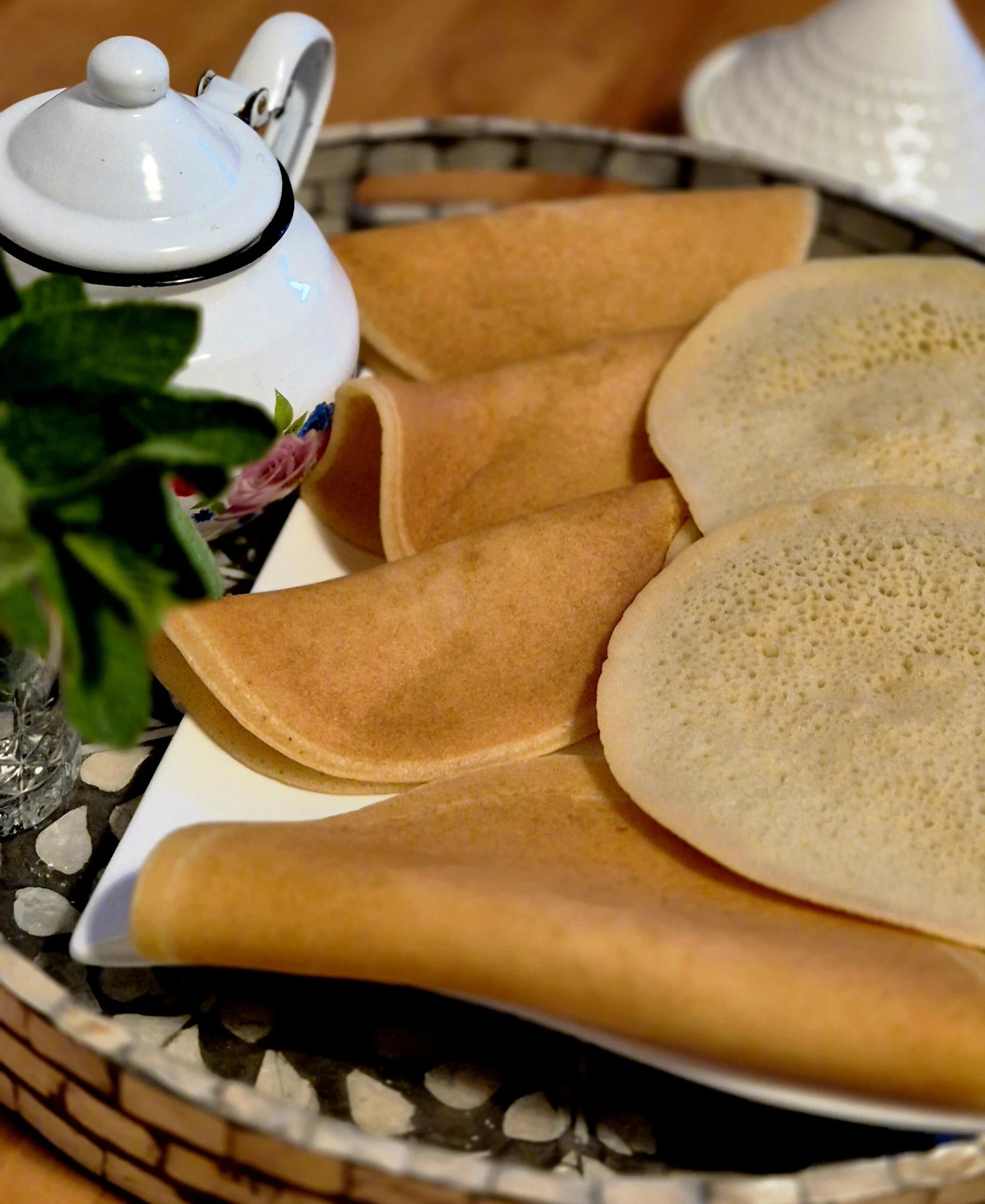 A serving plate with several round Moroccan pancakes, accompanied by a teapot and fresh mint leaves on a decorative tray