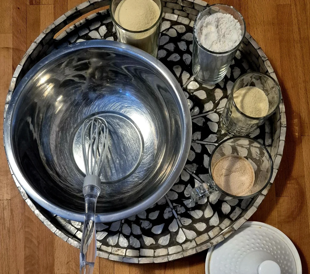 Moroccan Pancakes "Beghrir": A metal mixing bowl with a whisk on a decorative tray, surrounded by glasses of semolina flour, all-purpose flour, yeast, and sugar.