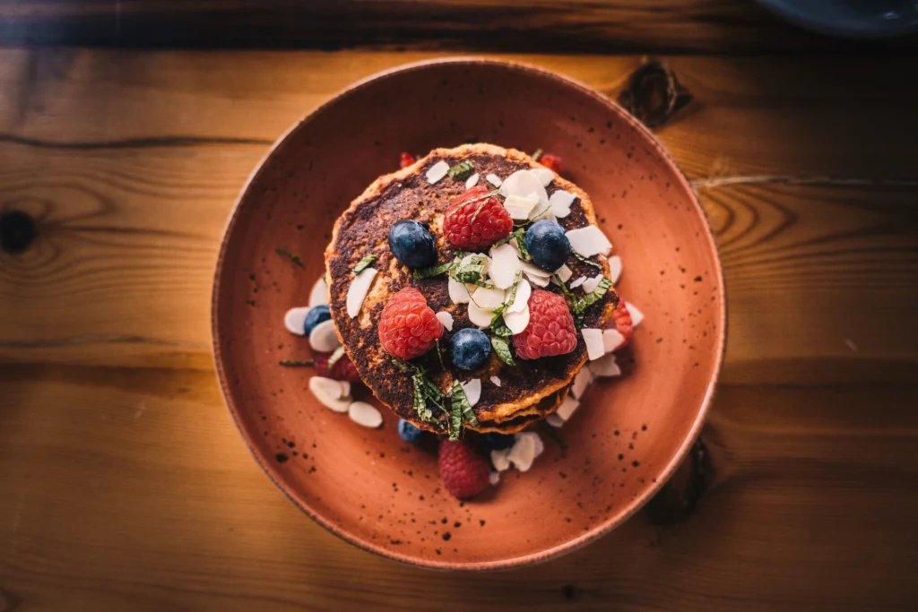 Stack of mini pancakes topped with fresh raspberries, blueberries, almond slices, and mint leaves on a rustic brown plate.