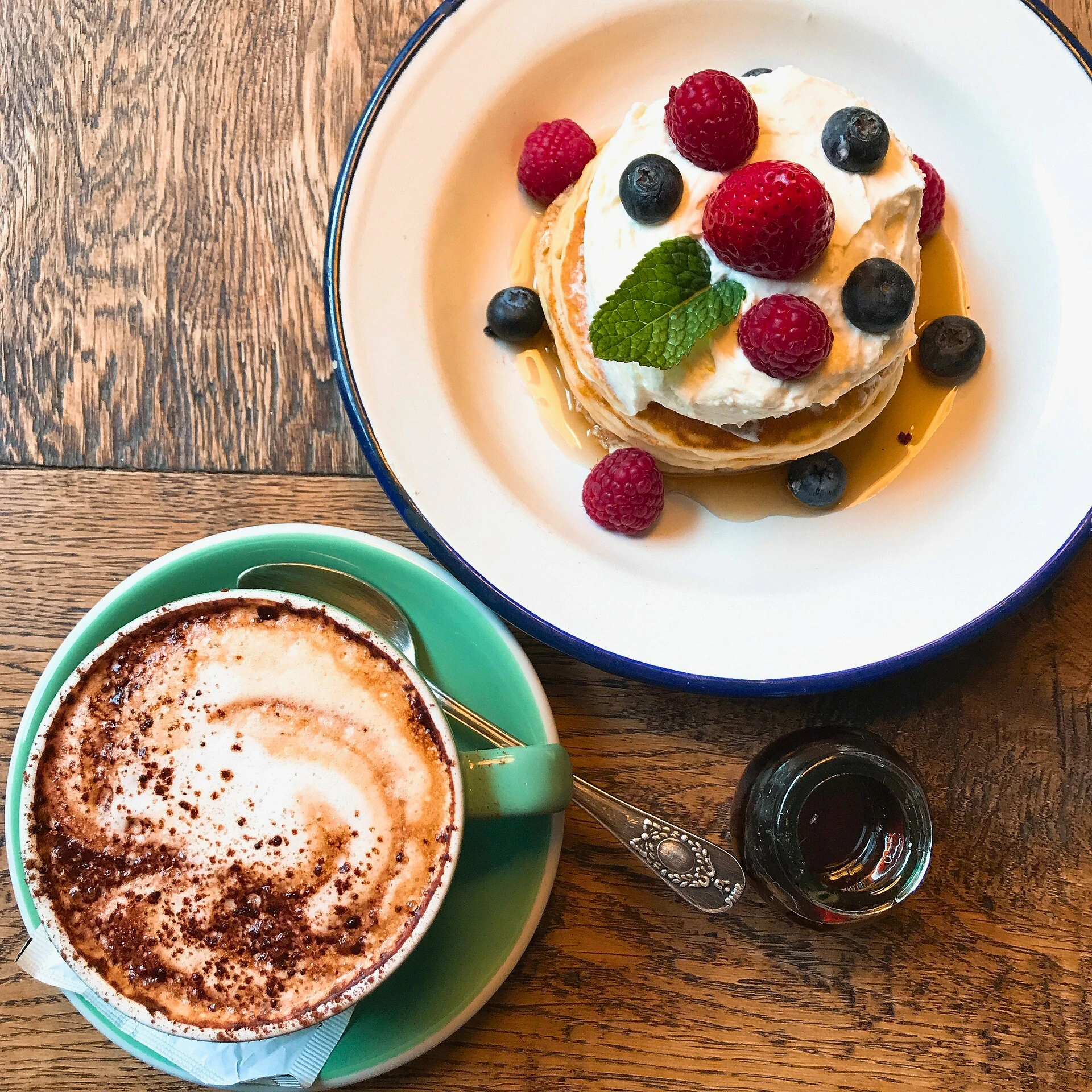 Mini pancakes with whipped cream, fresh berries, and syrup served alongside a cappuccino.