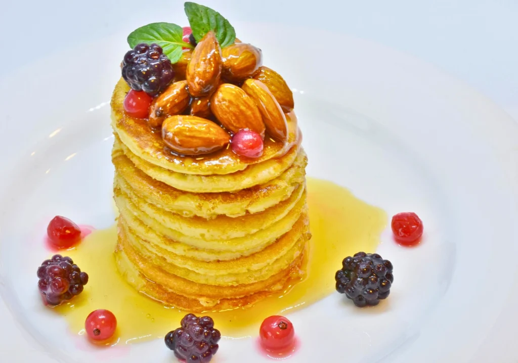 A tall stack of mini pancakes topped with glazed almonds, blackberries, and syrup on a white plate.